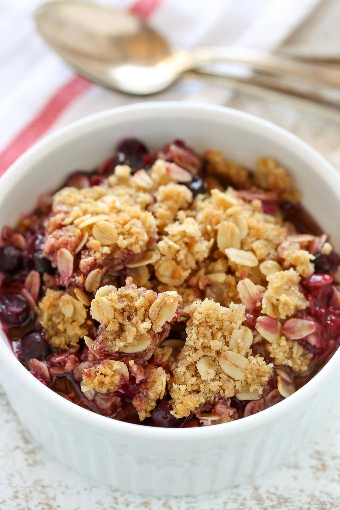 An individual serving of frozen berry crisp in a white ramekin. Two spoons and a towel rest in the background. 