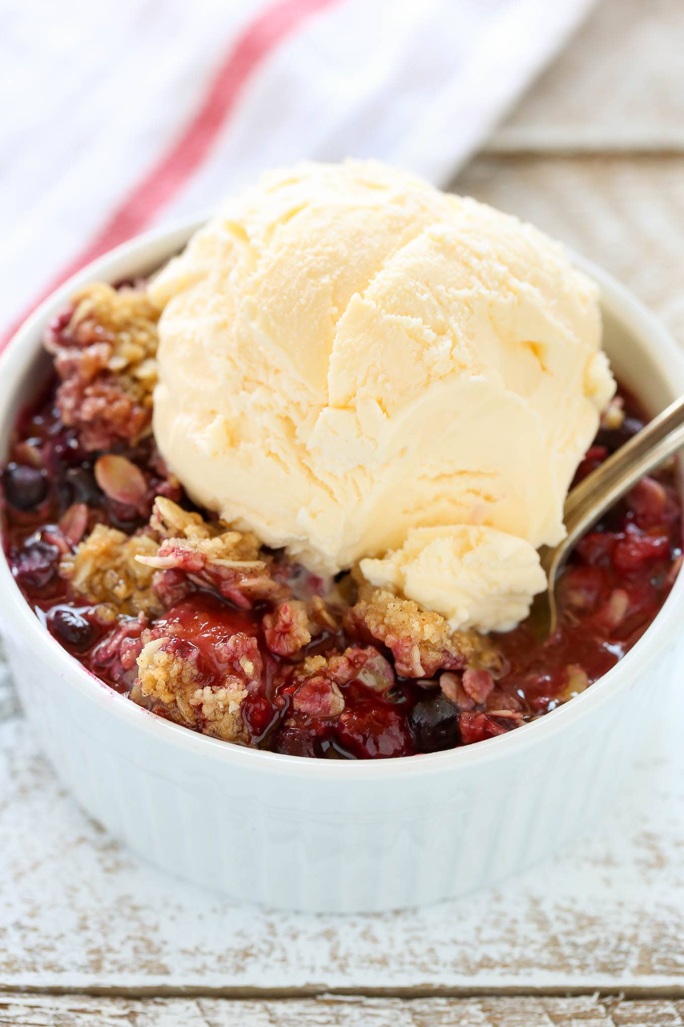 A white ramekin filled with mixed berry crisp with frozen berries, topped with a scoop of vanilla ice cream. 