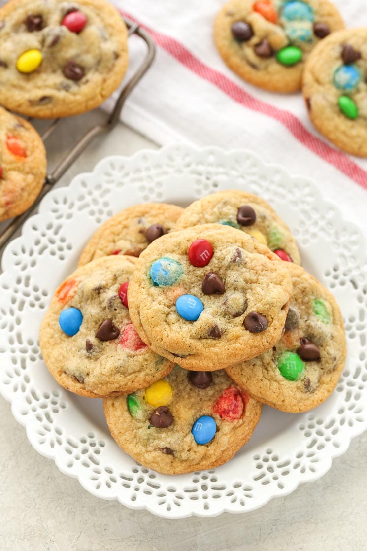 A white plate piled high with chocolate chip M&M's cookies. Additional cookies rest in the background. 