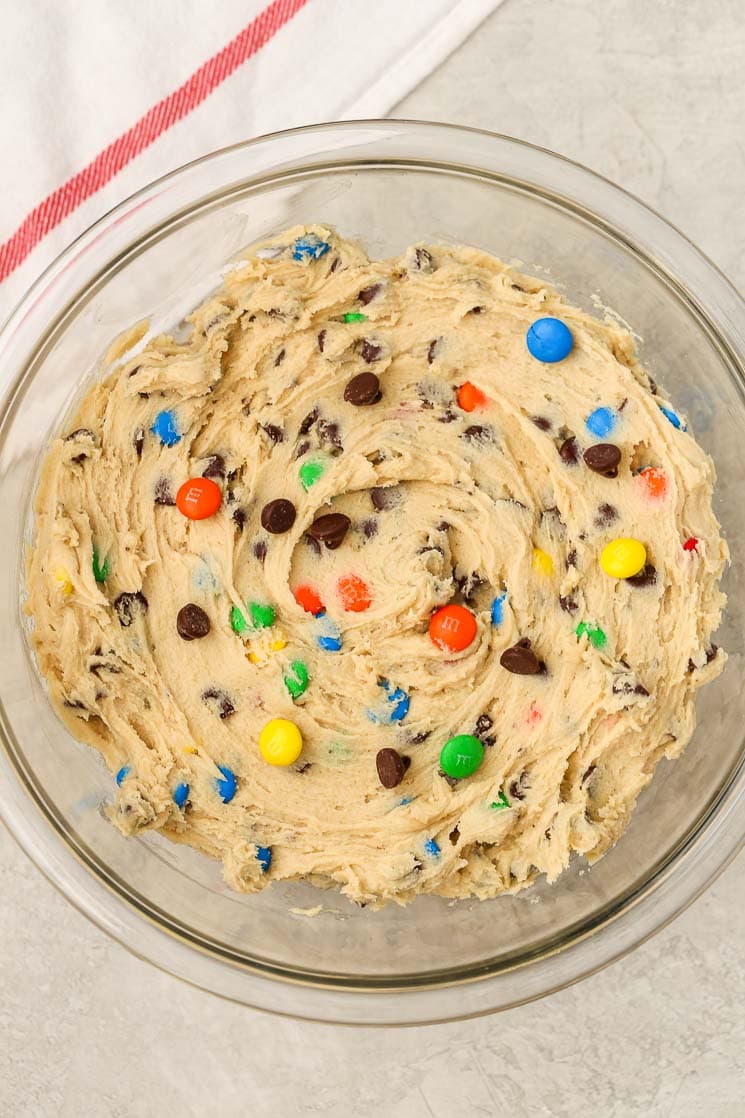 Glass mixing bowl filled with cookie dough that's studded with M&M's and chocolate chips. 