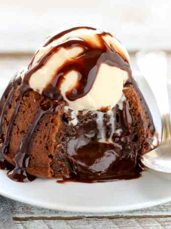 A molten chocolate cake topped with ice cream and chocolate sauce on a white plate. The cake has been cut into and the filling is oozing out.