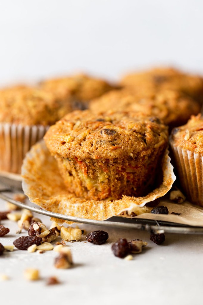 Side view of raisin apple carrot muffins on a wire cooling rack. The front muffin has had the paper liner pulled down. 