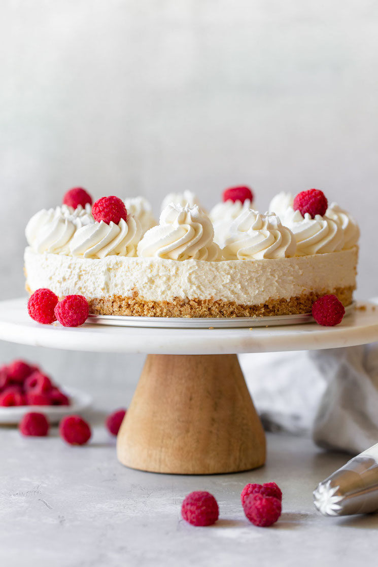 A whole no-bake cheesecake resting on a marble cake stand topped with whipped cream and raspberries.