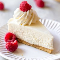 A single slice of no-bake cheesecake on a white plate topped with whipped cream and fresh raspberries.