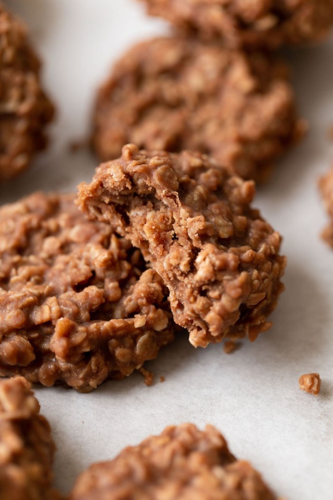 Several no-bake cookies on a piece of parchment paper. A cookie with a bite taken out of it is leaning on top of another cookie.