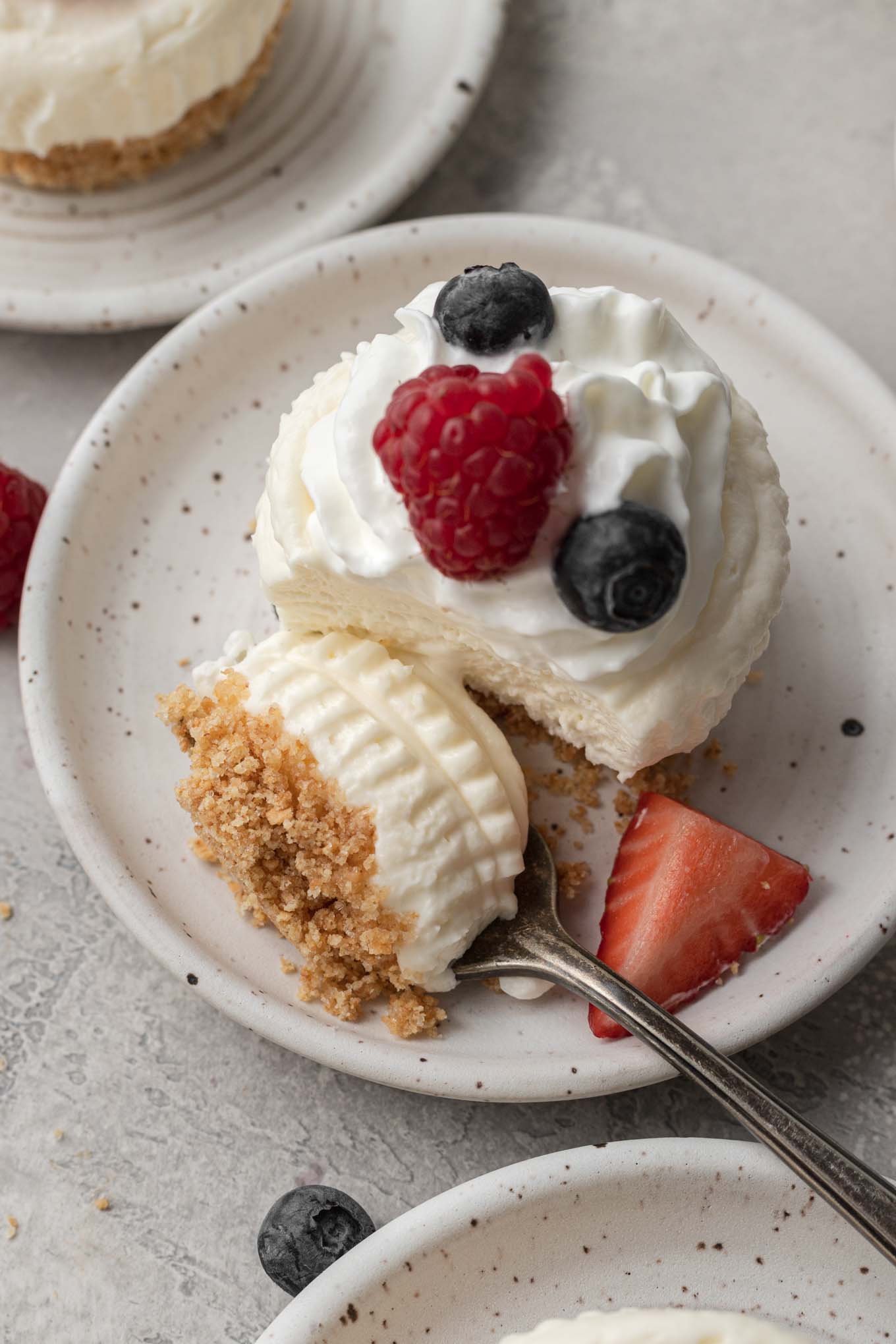 An overhead view of an individual no-bake cheesecake topped with whipped cream and fresh berries. A bite has been speared onto a fork. 