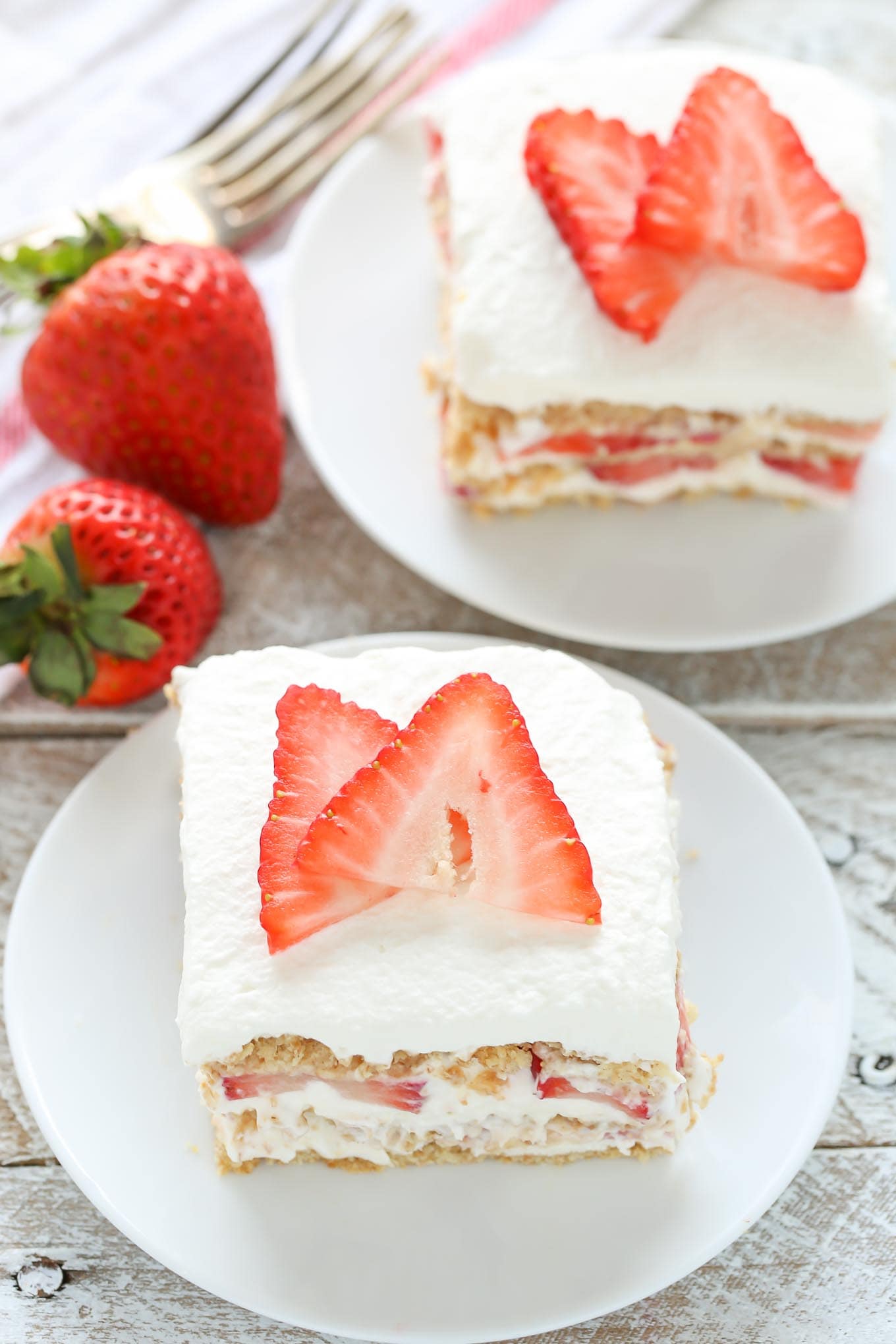 Two slices of strawberry icebox cake on white dessert plates. Two fresh berries and a fork rest on the side. 