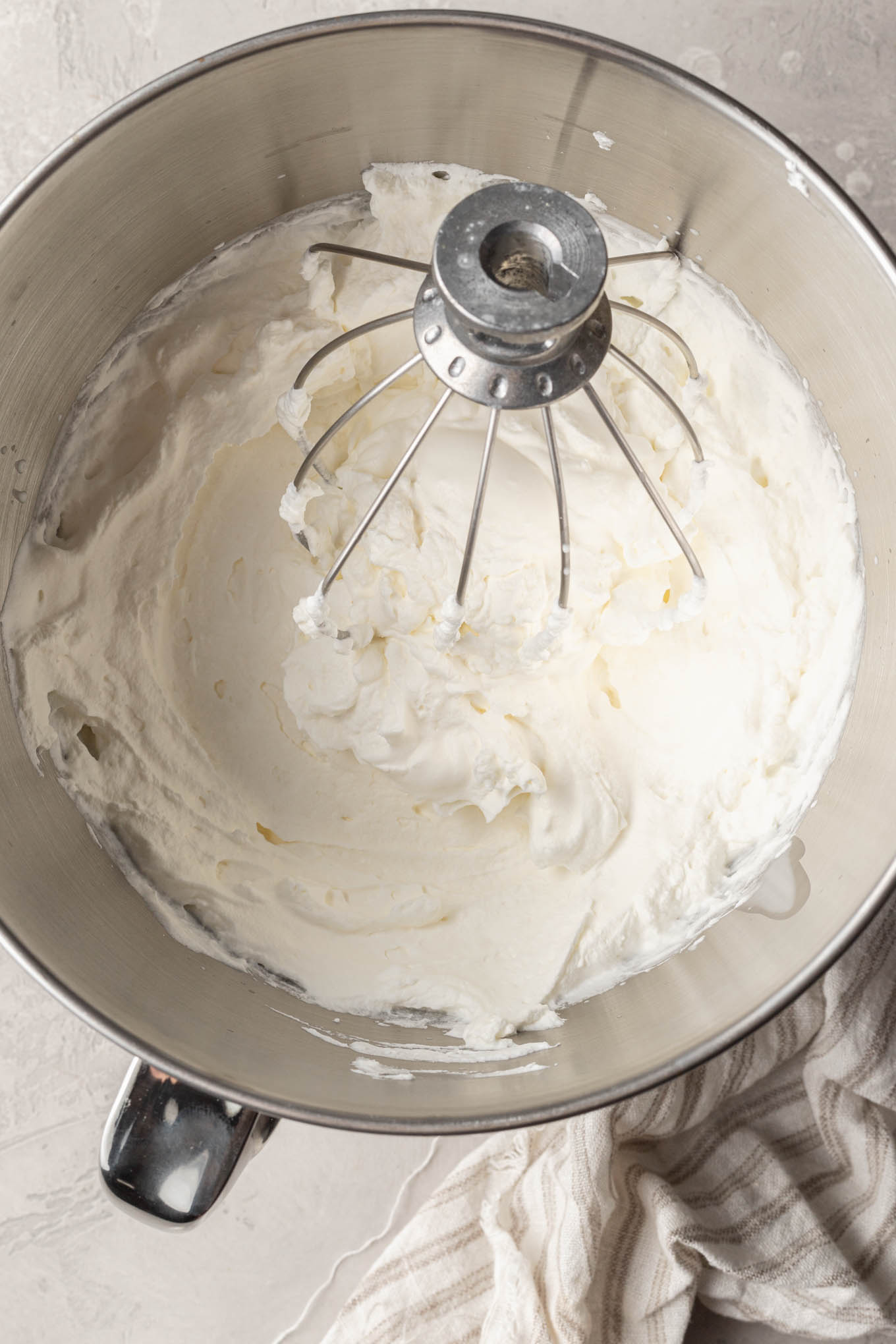 An overhead view of whipped cream beat to stiff peaks in the bowl of a stand mixer.