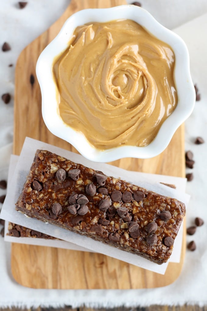 Overhead view of a stack of healthy peanut butter chocolate chip granola bars and a bowl of peanut butter on a wood board. 