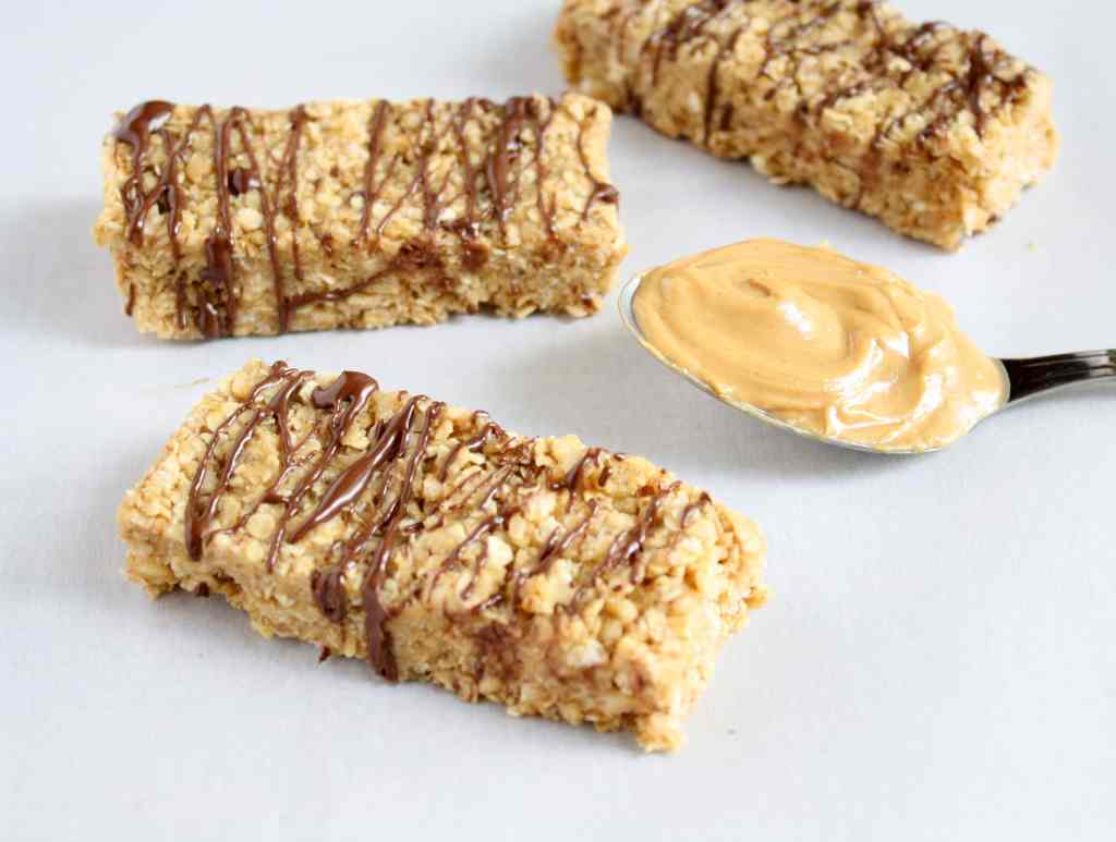 Three no bake peanut butter granola bars drizzled with chocolate. A spoonful of peanut butter rests nearby. 