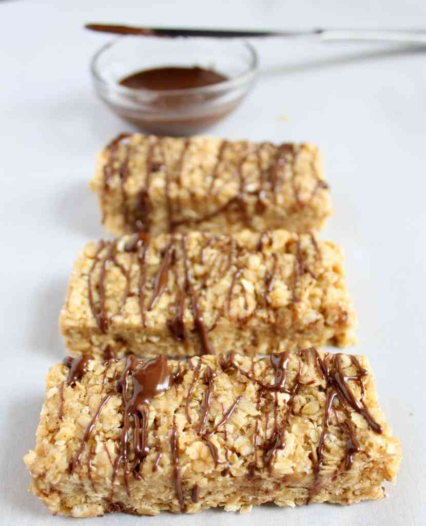 A row of three chewy peanut butter granola bars drizzled with chocolate. A bowl of melted chocolate is in the background. 