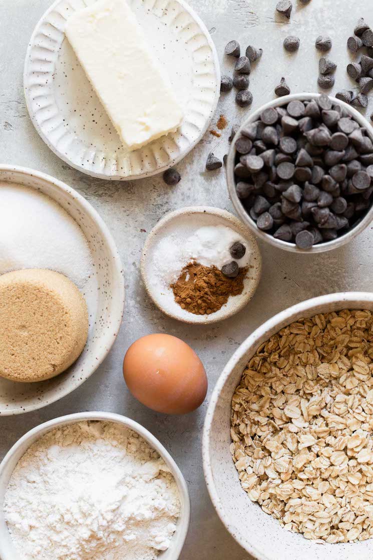 The ingredients needed to make oatmeal chocolate chip cookies separated onto speckled bowls and plates.