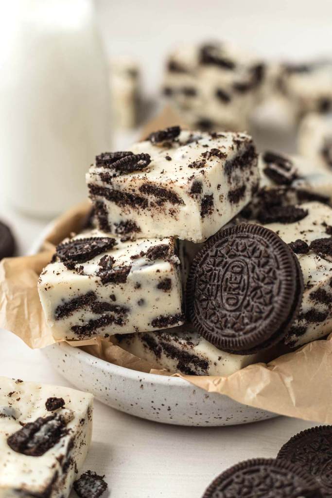 Slices of Oreo fudge in a white speckled bowl. A glass milk jug rests in the background. 