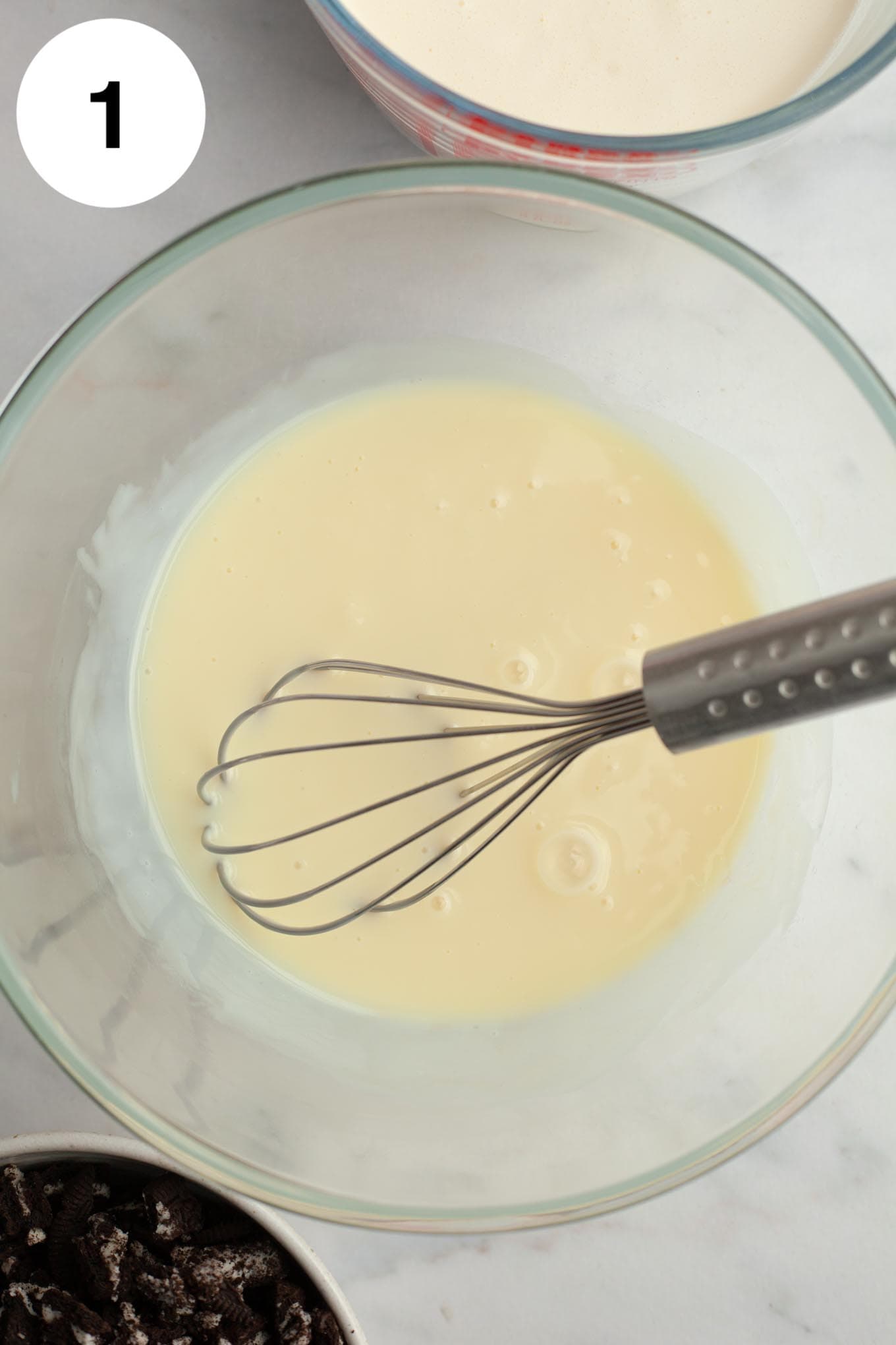 An overhead view of sweetened condensed milk in a glass mixing bowl with a whisk. 