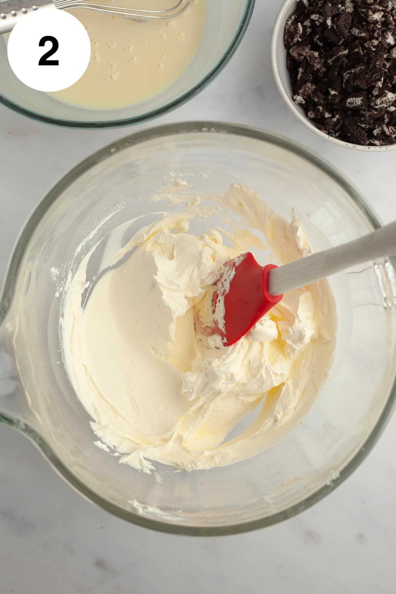 An overhead view of whipped cream in a glass mixing bowl with a rubber spatula. 