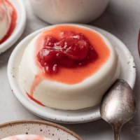 Panna cotta topped with homemade strawberry sauce on a speckled white plate. A spoon rests next to it.