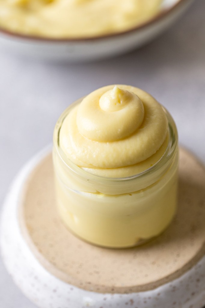 A small glass jar filled with vanilla pastry cream. The jar rests on an overturned bowl. More pastry cream rests in the background. 