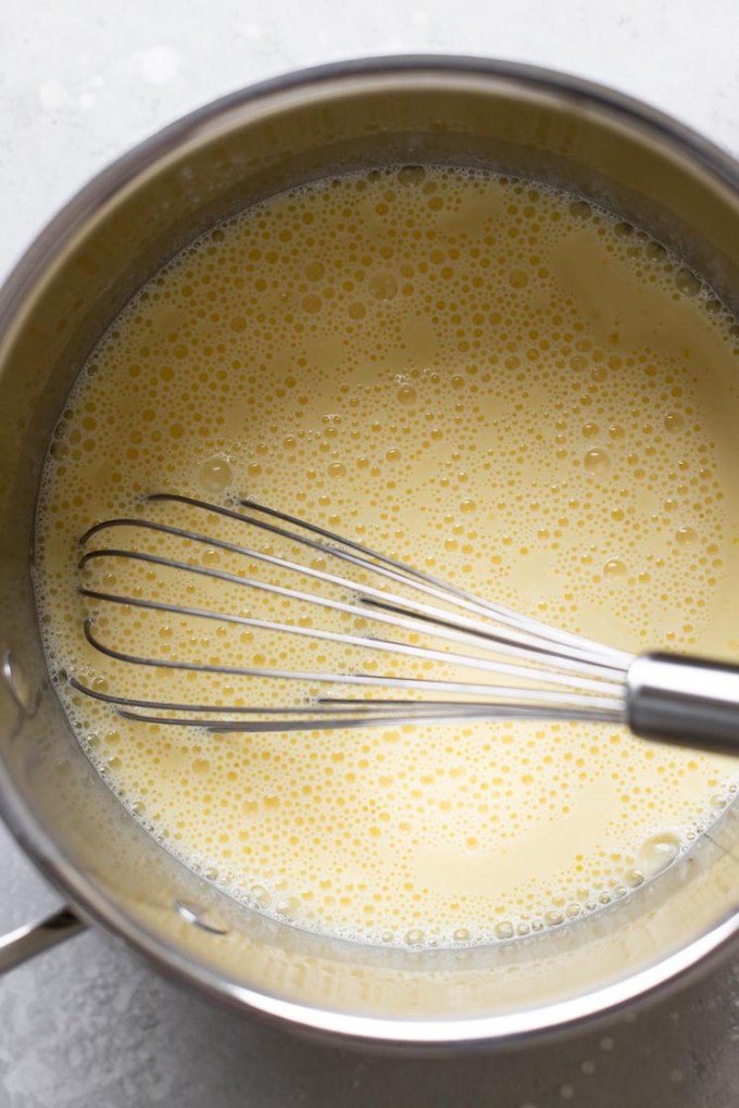 Overhead view of a saucepan filled with milk, egg yolks, cornstarch, and sugar. A whisk rests inside the pan.