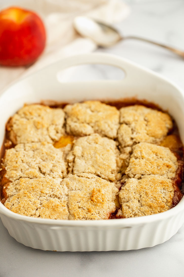 A peach cobbler fresh out of the oven with an antique serving spoon and a peach in the background. 