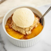 A white ramekin on a marble surface filled with peach cobbler and a scoop of ice cream on top.