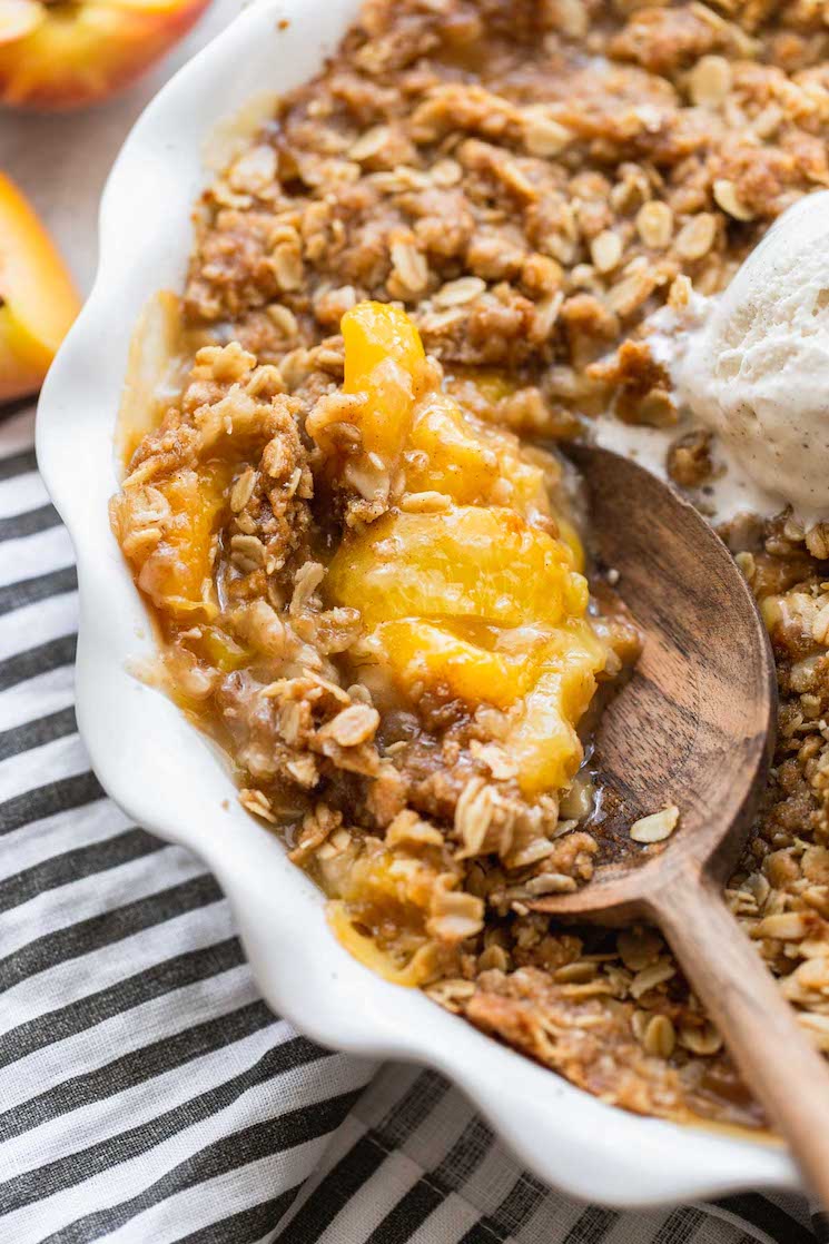 A finished peach crisp in a round baking dish being scooped with a wooden spoon.