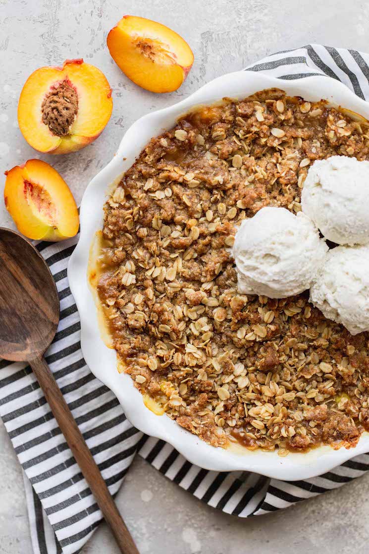 A finished peach crisp in a white baking dish topped with ice cream and another a cut peach beside it.