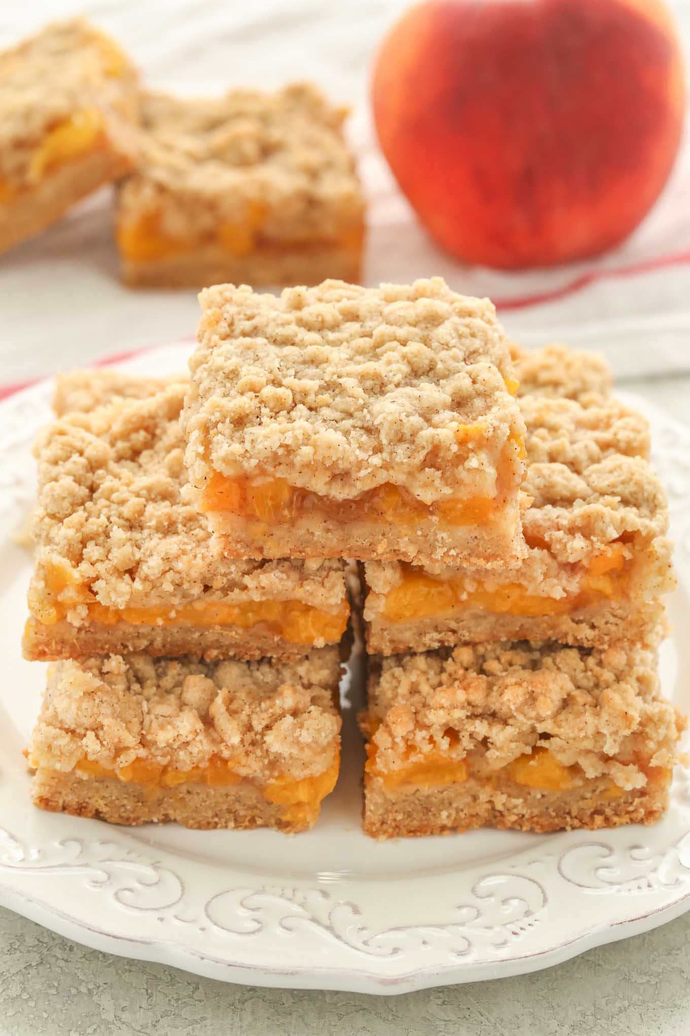 A pile of peach crumble bars on a white plate. A fresh peach and two more bars rest in the background. 