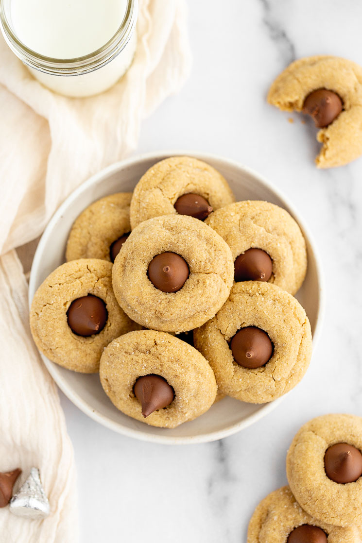 A plate with several peanut butter blossom cookies stacked on it with a glass of milk and a cookie with a bite taken out of it in the background.