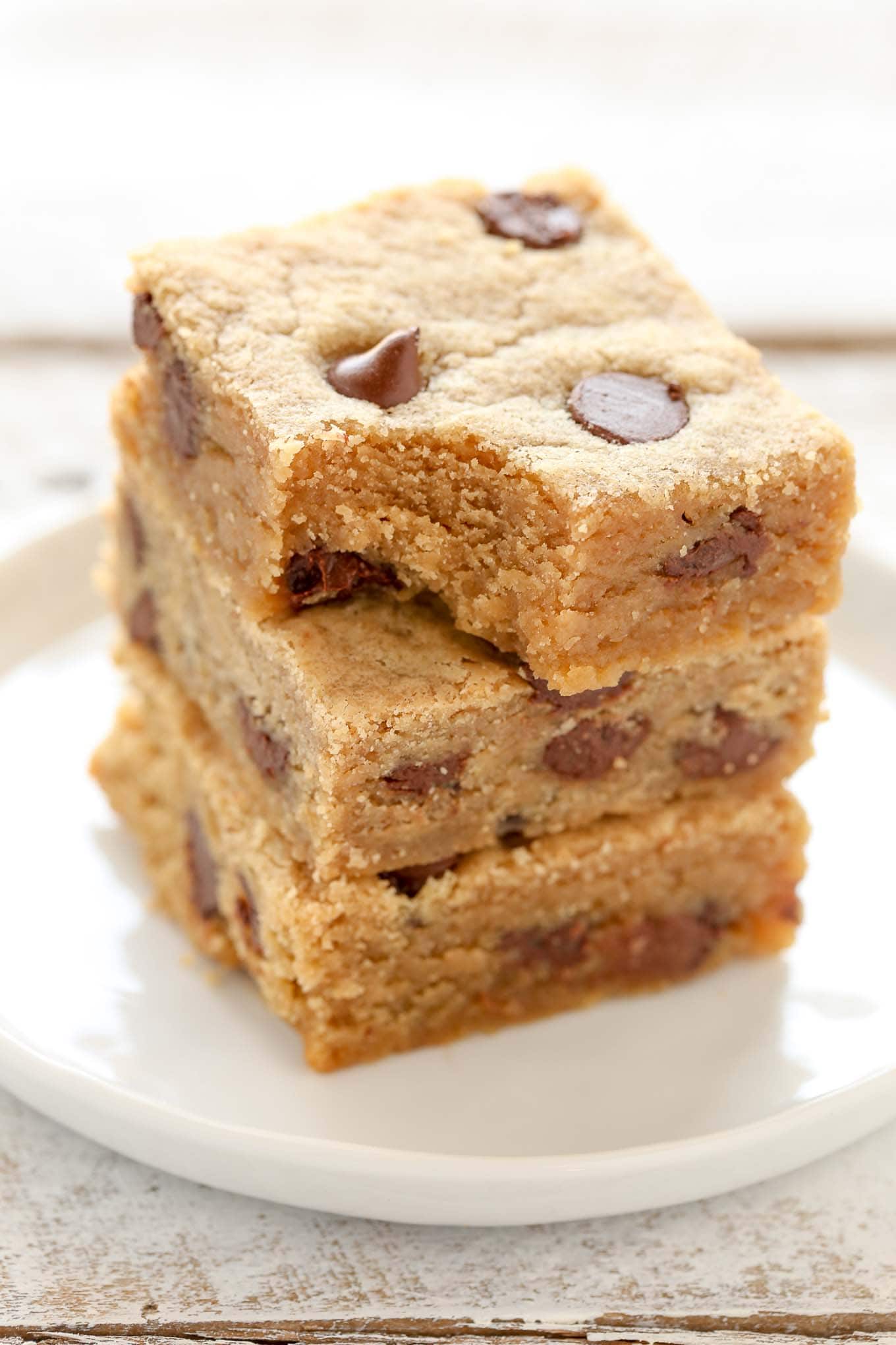 A stack of three peanut butter chocolate chip bars on a white plate. The top bar has a bite missing. 