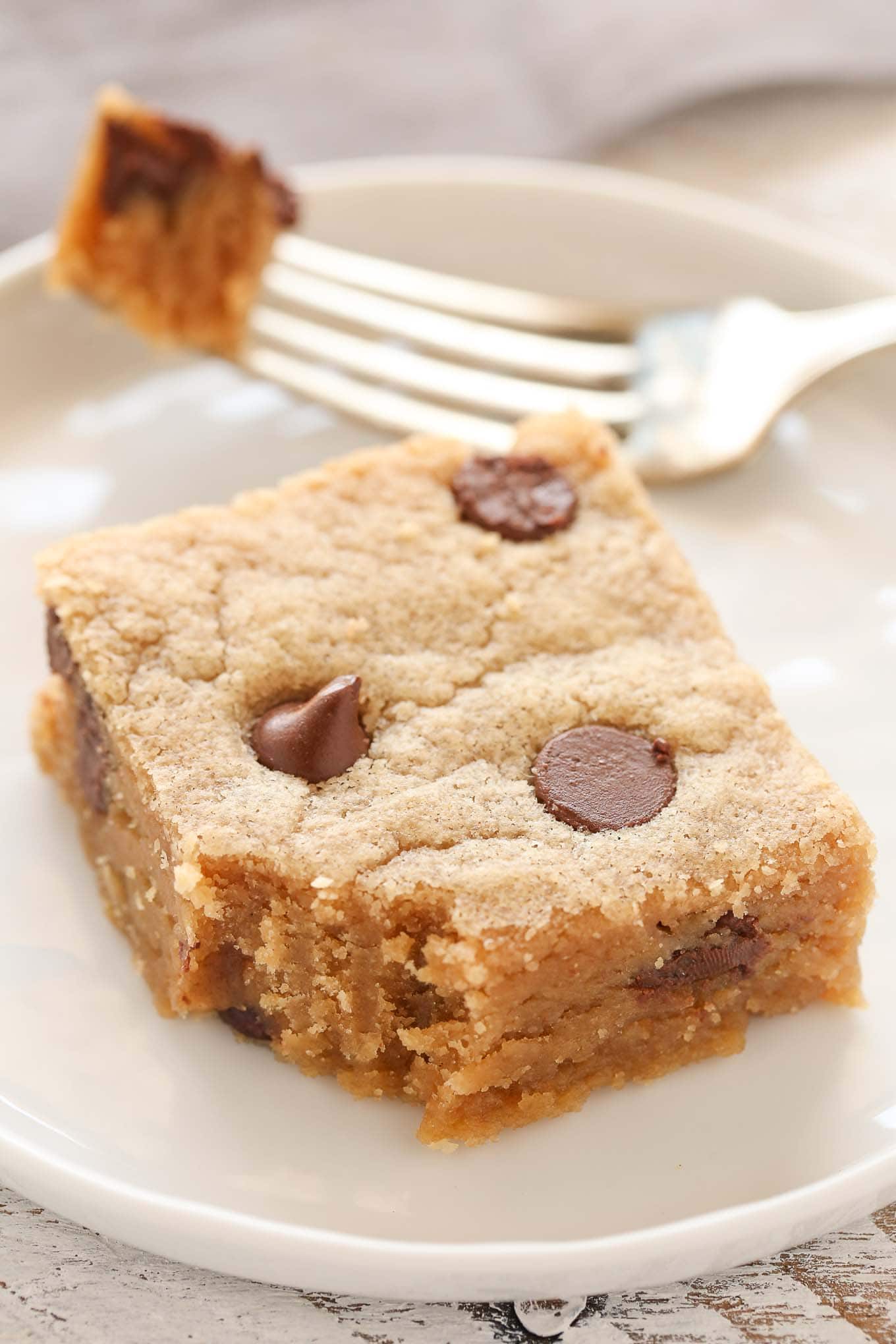 A peanut butter chocolate chip cookie bar on a white plate with a fork. A bite has been taken from the bar. 