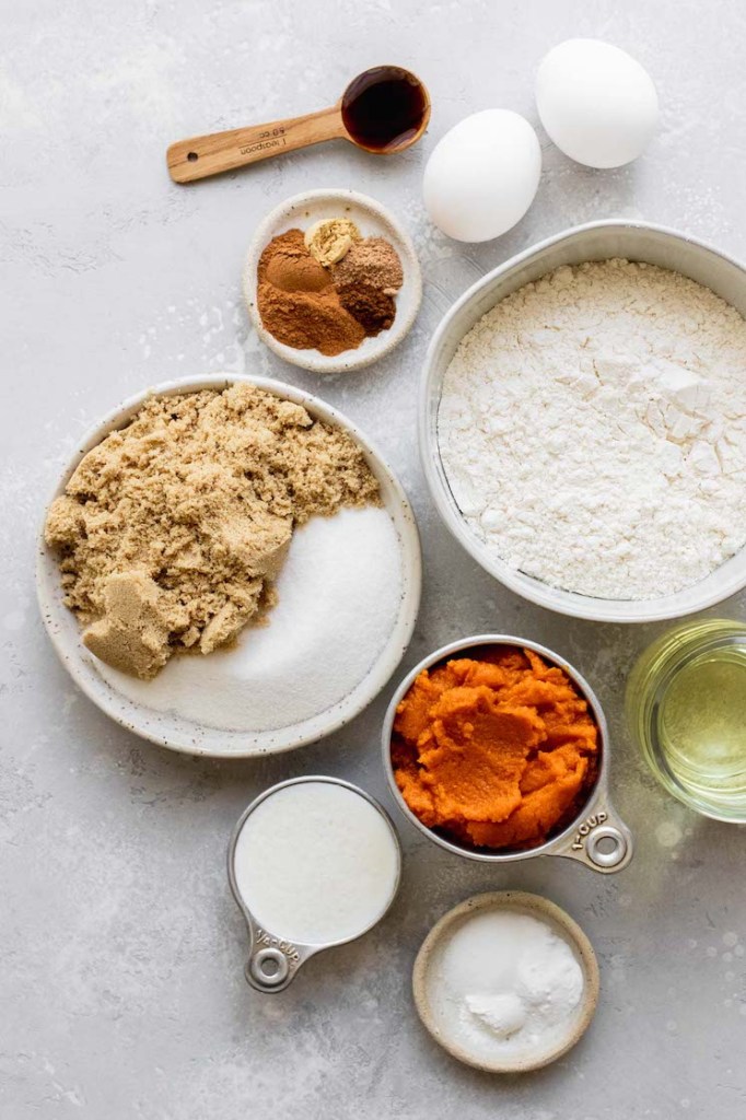 The ingredients needed to make pumpkin bread displayed in bowls and sitting on top of a rustic gray surface.