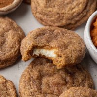 Pumpkin cheesecake cookies on a gray surface. One cookie has a bite missing to show the cream cheese center.