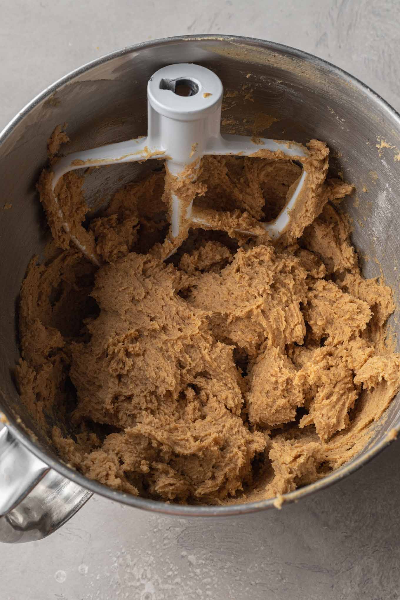 An overhead view of pumpkin cookie dough in the bowl of a stand mixer, with the paddle attachment in the bowl. 