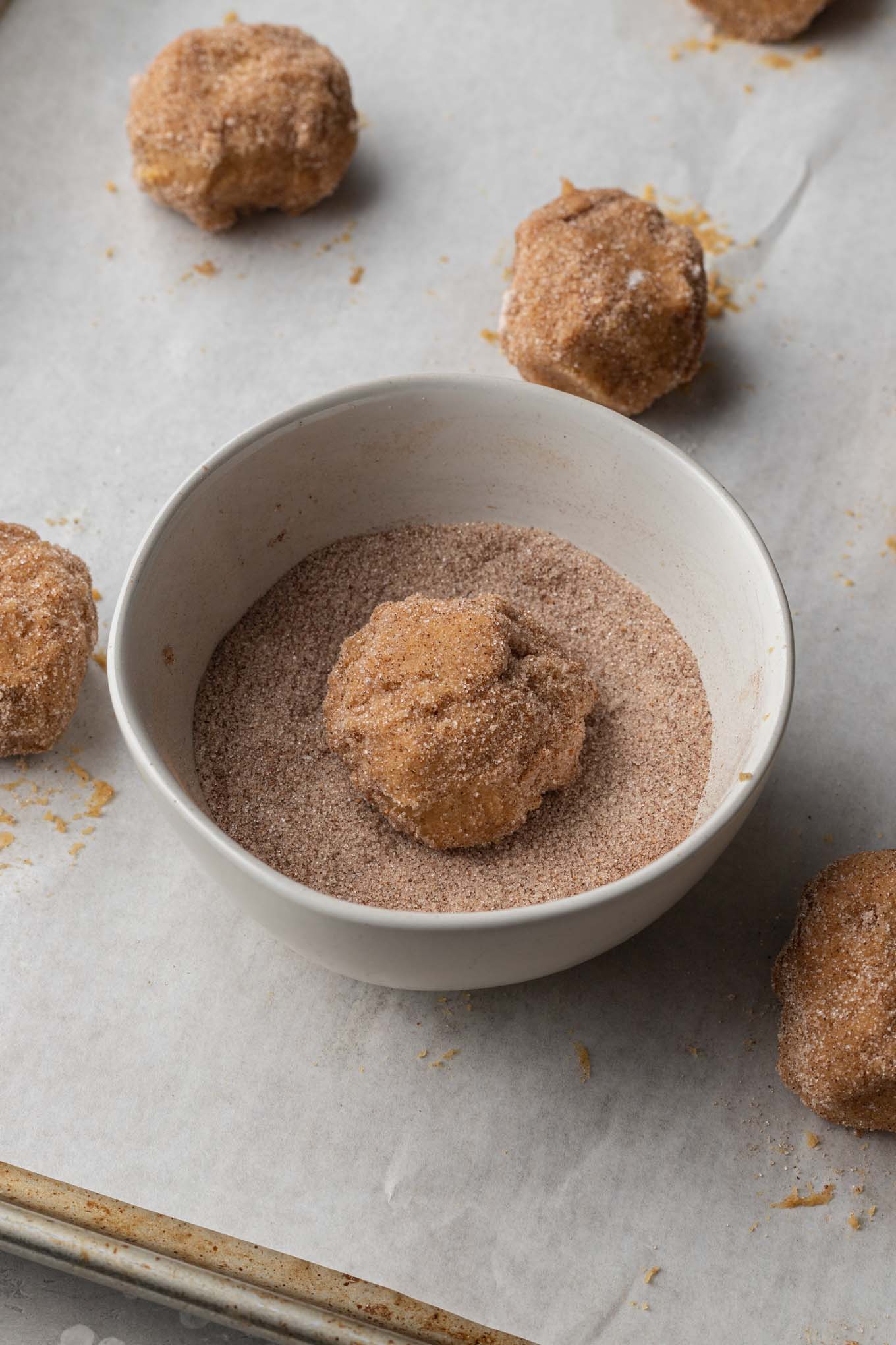 A bowl of cinnamon sugar with a cookie dough ball resting in the middle. The bowl is on a parchment-paper lined cookie sheet. 