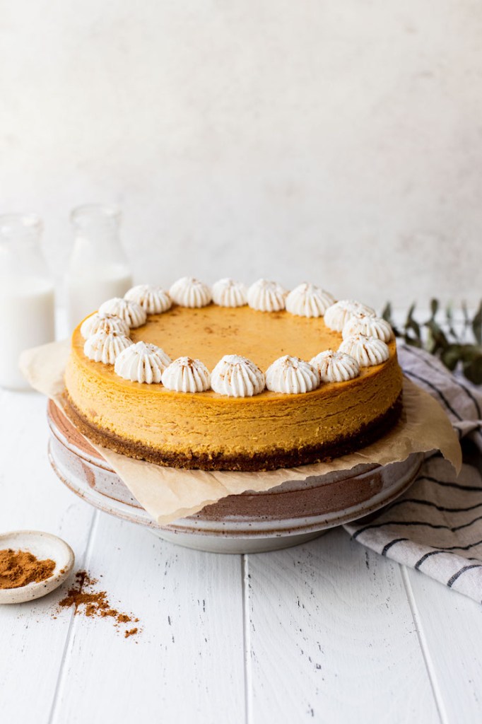 A whole pumpkin cheesecake on a cake stand with milk and cinnamon around it.