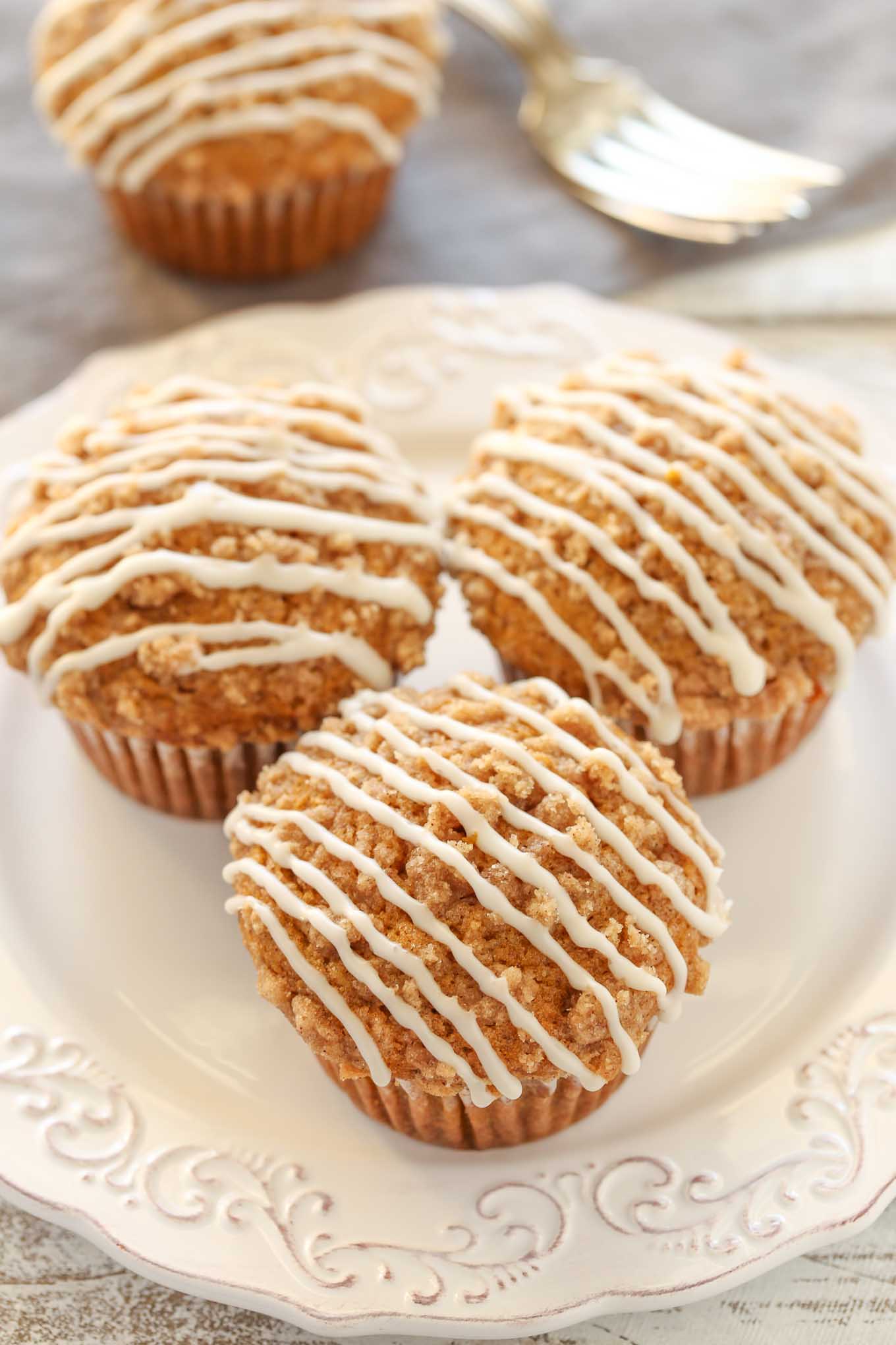 Three glazed pumpkin streusel muffins on a white plate. Another muffin and a fork rest in the background. 