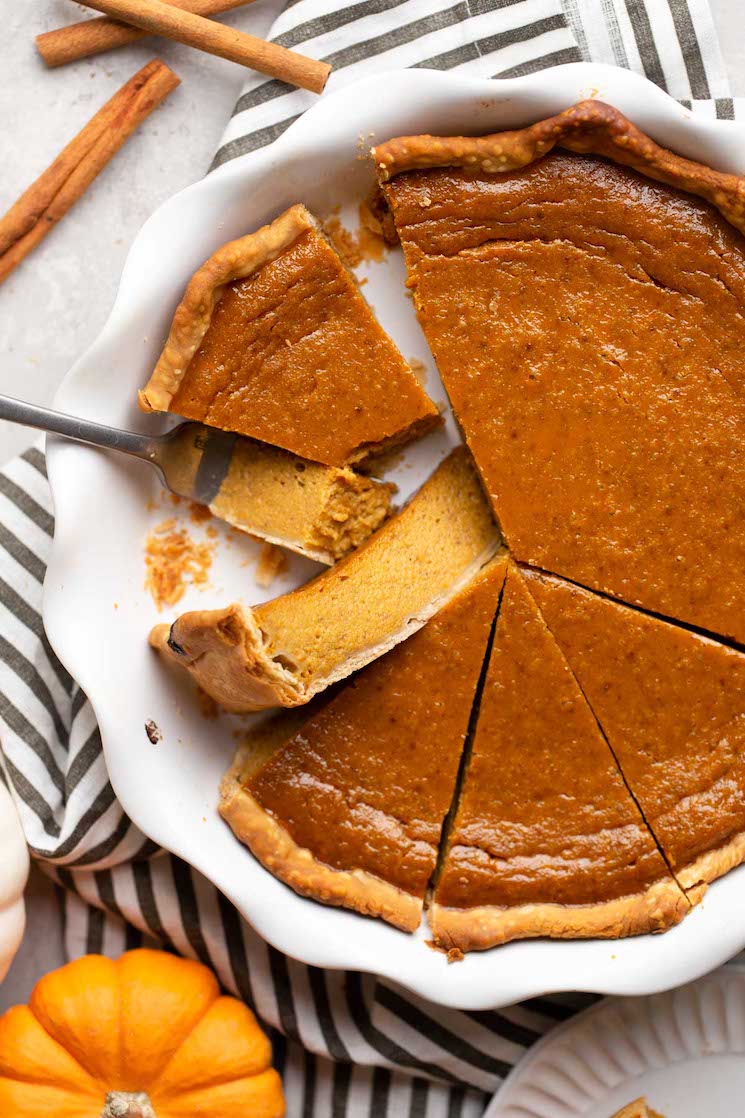 An overhead view of a pumpkin pie from scratch that's been cut into slices. Cinnamon sticks and a mini pumpkin rest nearby. 