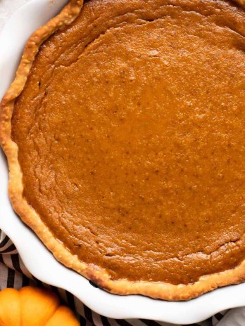 An overhead view of a homemade pumpkin pie in a white pie plate. A mini pumpkin and cinnamon sticks are near it.