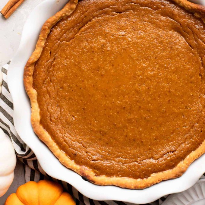 An overhead view of a homemade pumpkin pie in a white pie plate. A mini pumpkin and cinnamon sticks are near it.