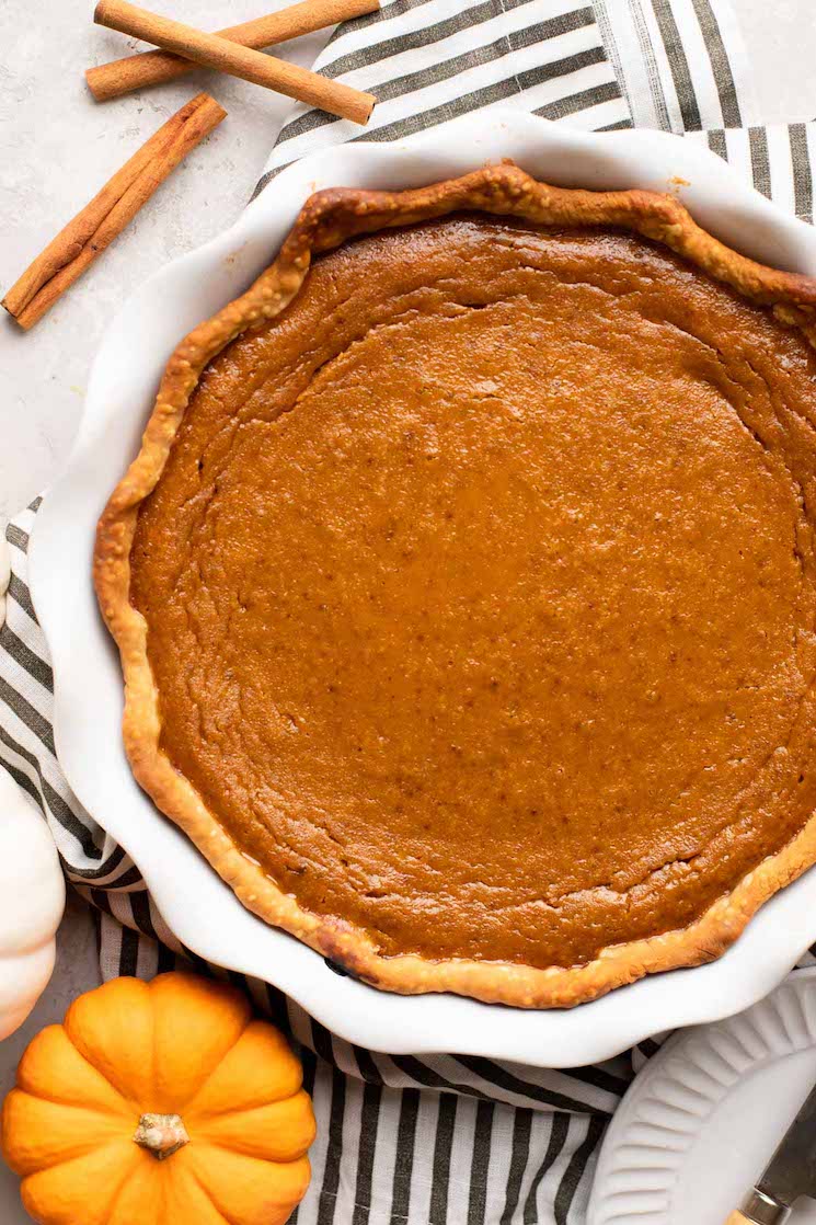 An overhead view of a homemade pumpkin pie in a white pie plate. A mini pumpkin and cinnamon sticks are near it. 