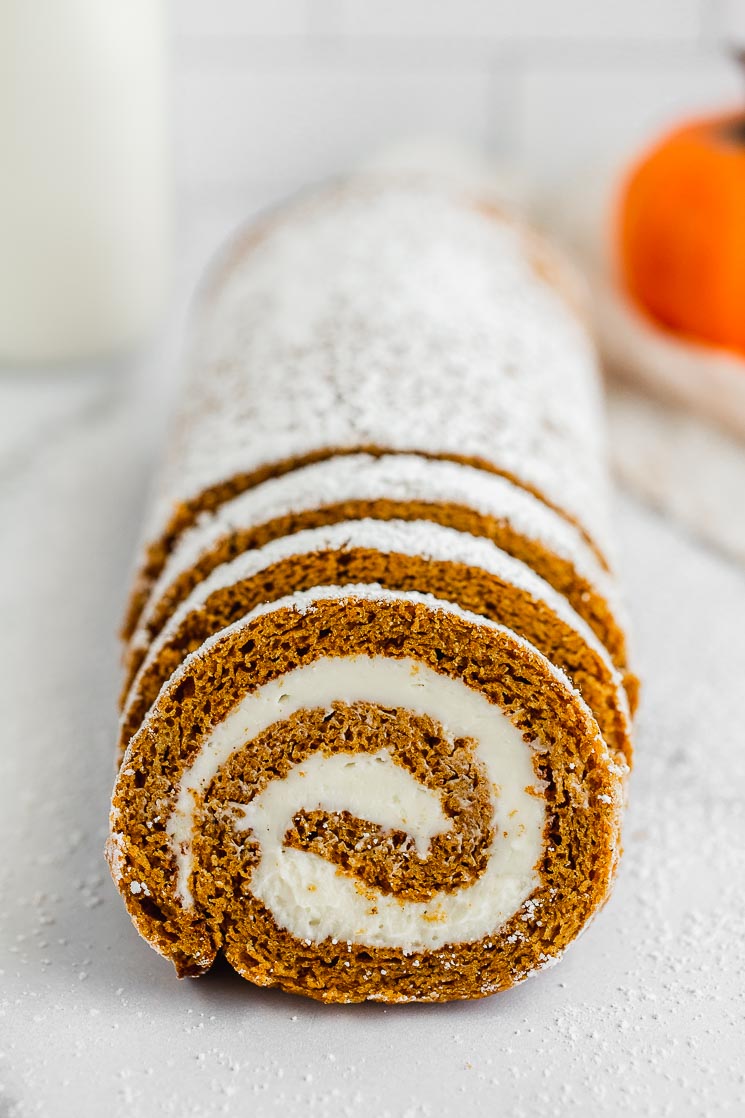 A sliced pumpkin roll with milk and a pumpkin in the background.