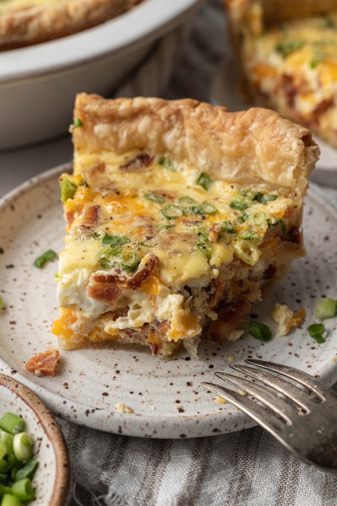 A slice of quiche with a bite missing, on a speckled plate. 