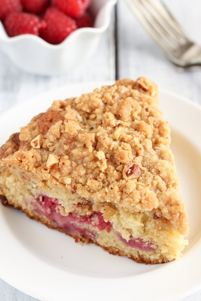 A slice of raspberry almond cake with crumb topping on a white plate. A bowl of fresh raspberries rest in the background. 