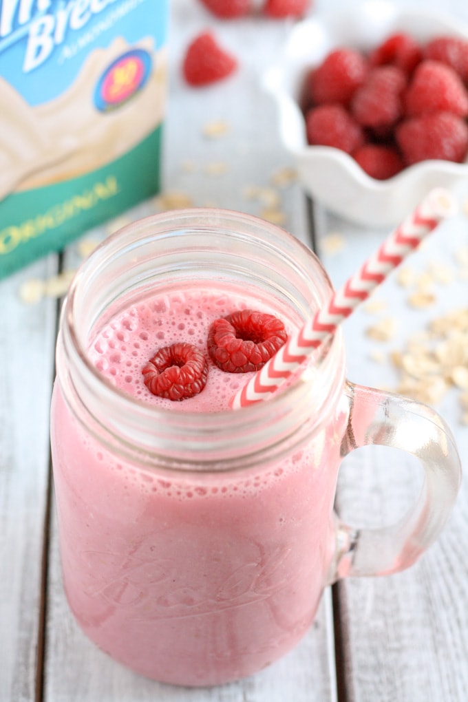 A raspberry smoothie in a glass mason jar with a striped straw. A carton of almond milk and a bowl of fresh raspberries rest in the background.