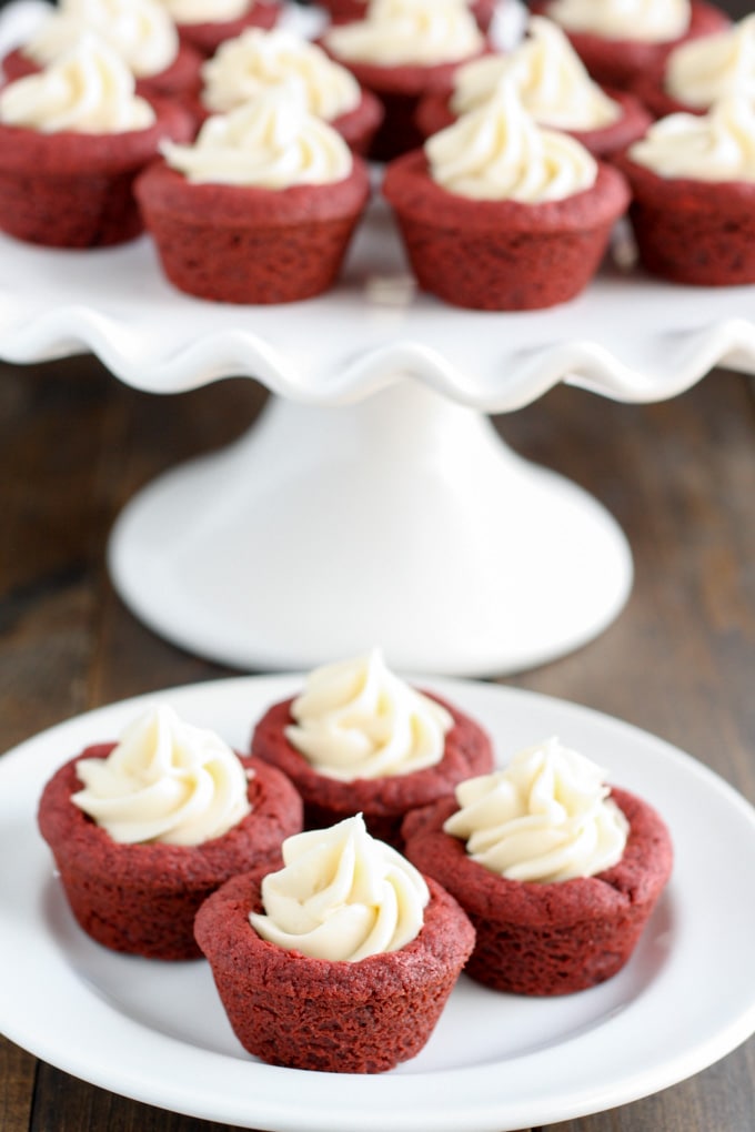 Deep dish red velvet cookie cups with cream cheese frosting on a white plate. A cake stand of cookie cups is in the background. 