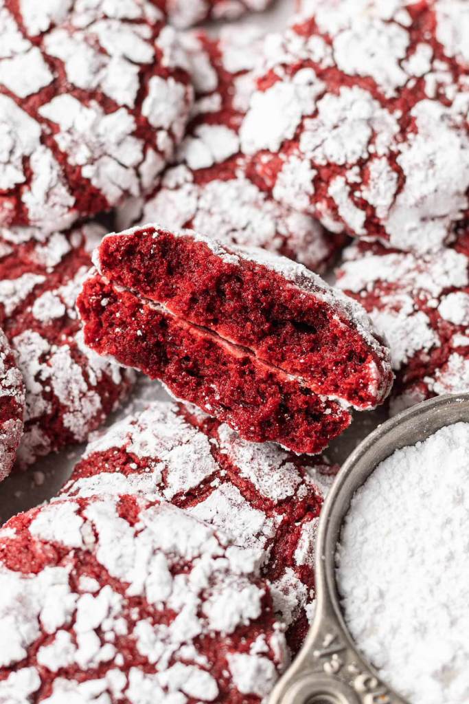 A close up view of a red velvet cookie that's been broken in half and stacked on itself, amidst a pile of cookies. 