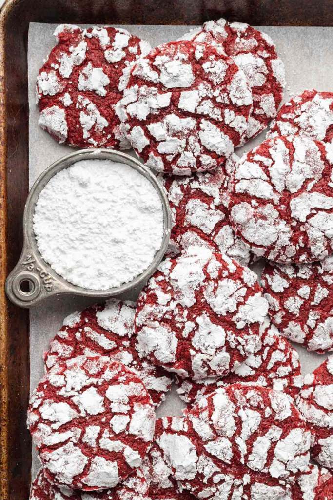 An overhead view of red velvet crinkle cookies piled on a baking sheet. A measuring cup of powdered sugar rests by the cookies. 