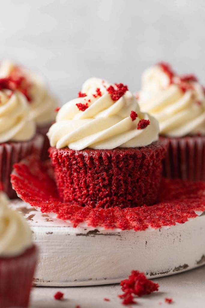 Three homemade red velvet cupcakes on a white platter. The front cupcake has had its liner pulled down. 