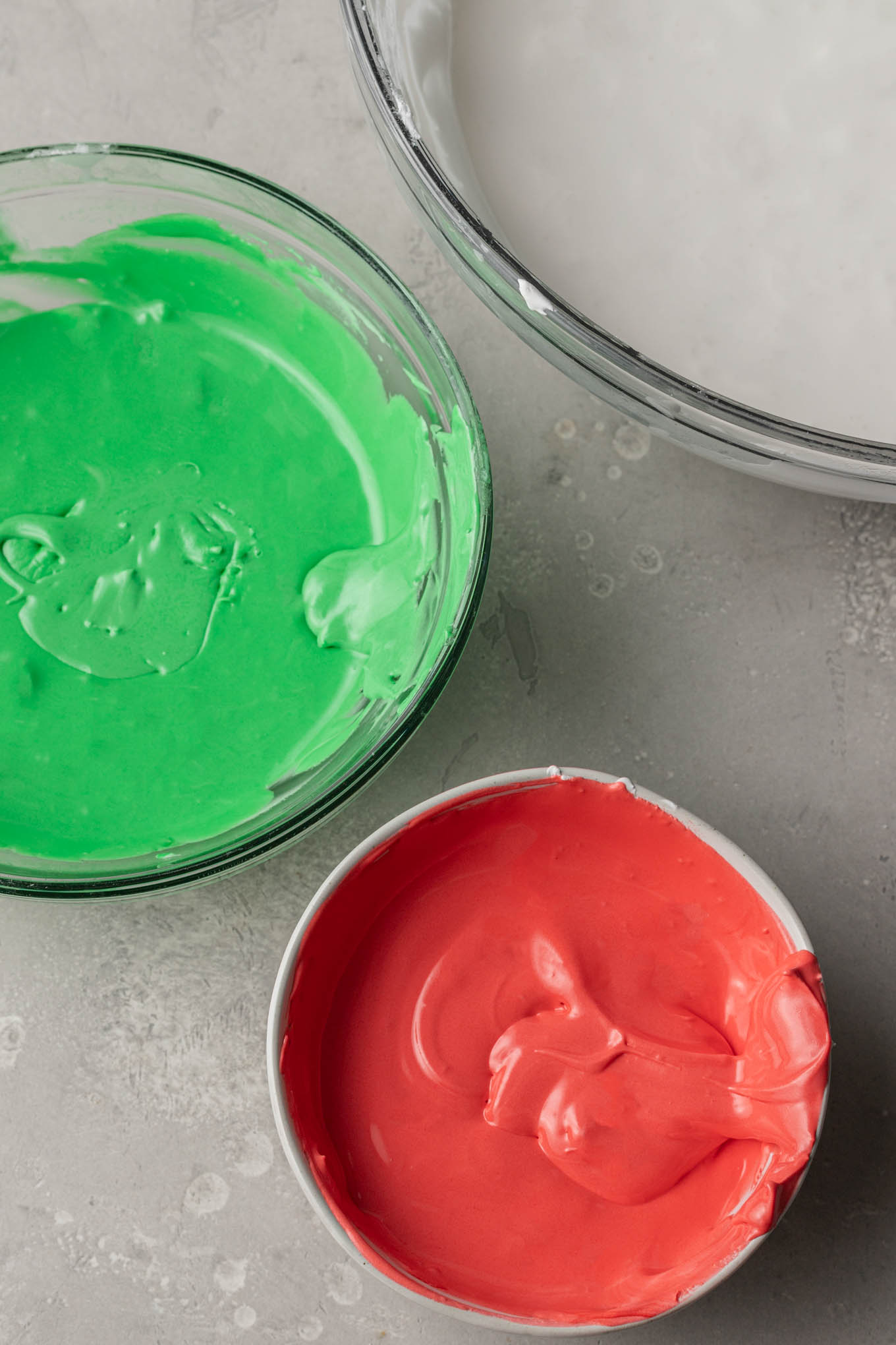 An overhead view of green, red, and white icing in mixing bowls.