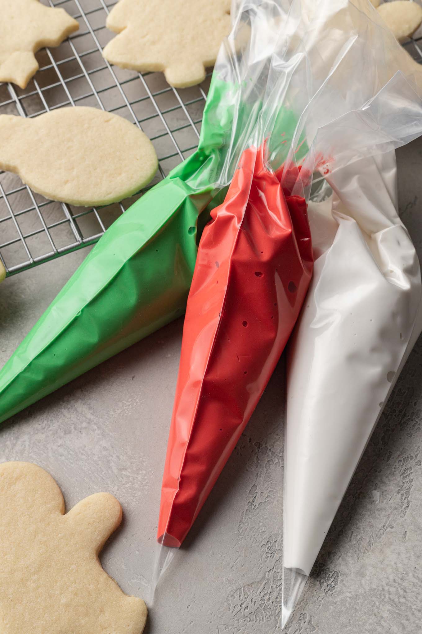 An overhead view of three piping bags filled with red, green, and white royal icing. A few sugar cookies are laying on a cooling rack by the piping bags.